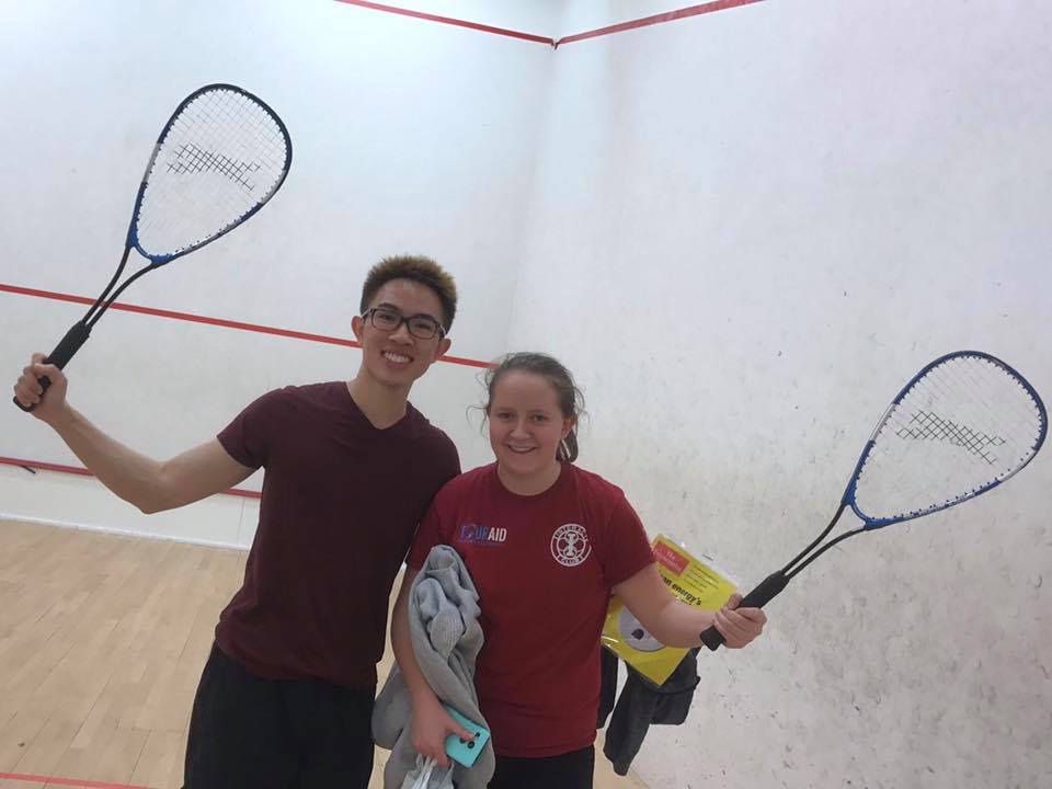 Ellie with a friend, both holding up their racquets and smiling, in the Christ's College squash court