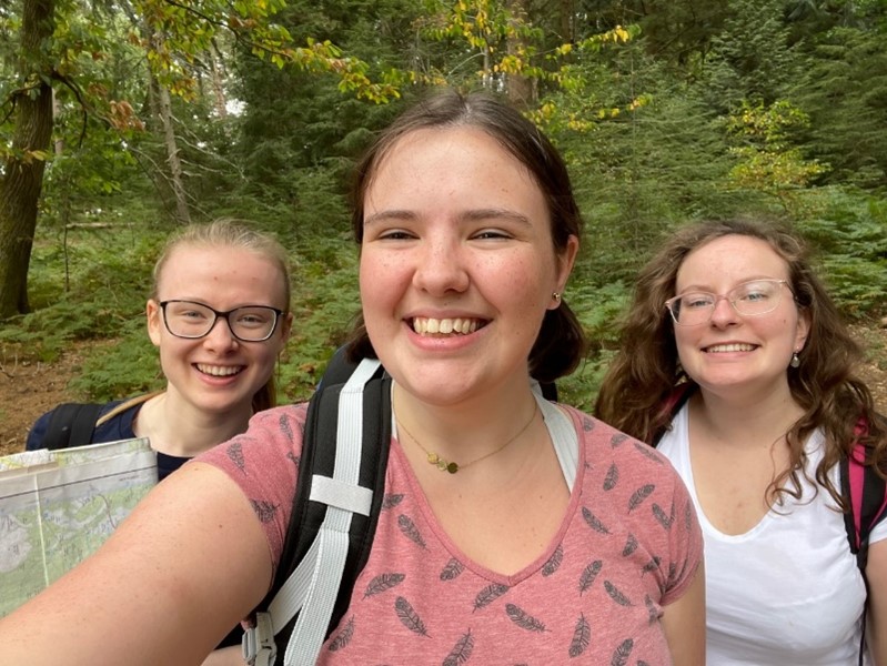 Selfie of three students, with Annie in the middle. They are in a forest, wearing rucksacks, and one of them holds a map.